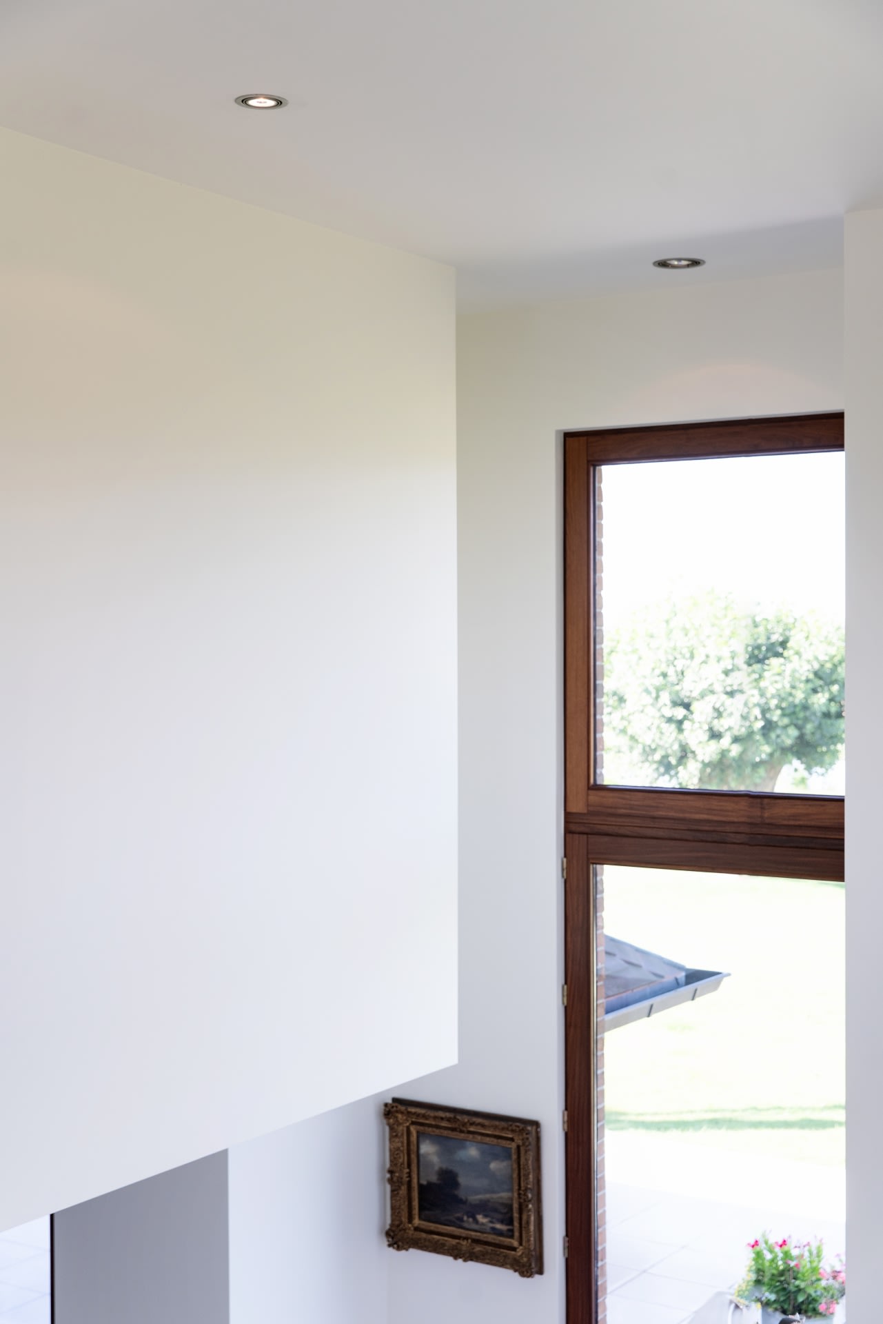 Intérieur moderne avec des murs blancs, une grande fenêtre avec une moulure en bois offrant une vue verdoyante sur l'extérieur, et un tableau encadré accroché au mur.
