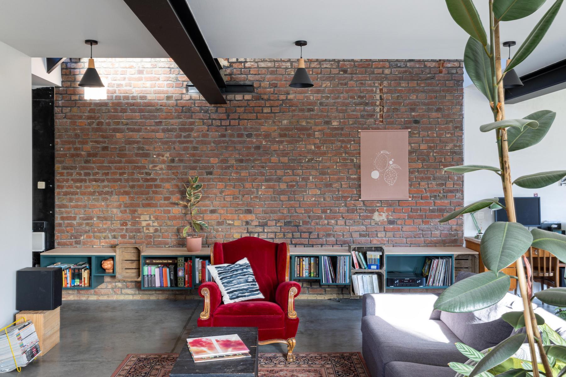 Un salon avec un fauteuil rouge, un canapé gris, une plante en pot, un mur en briques, des étagères avec des livres et des disques, et un tapis à motifs sur un sol en béton.