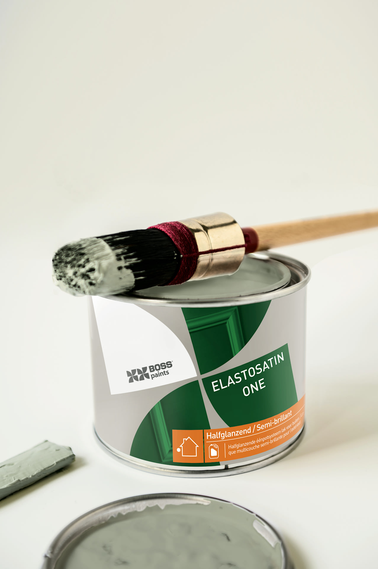 Un pinceau recouvert de peinture verte repose sur un pot de peinture BOSS ELASTOSATIN ONE ouvert, muni d'un couvercle, à côté duquel se trouve une tache de peinture verte sur une surface de couleur claire.