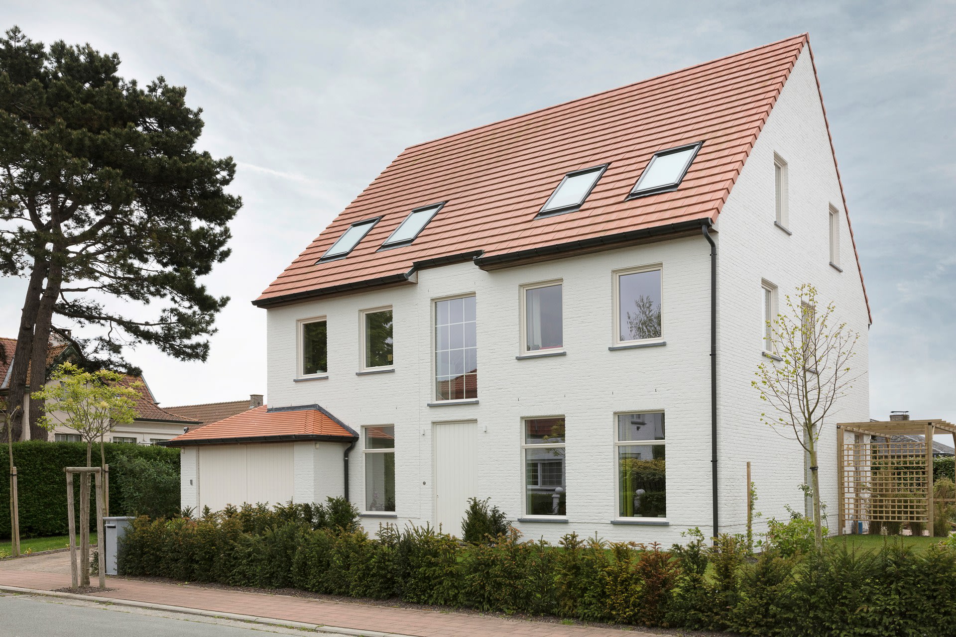 Een modern wit huis van twee verdiepingen met een rood pannendak, veel ramen en dakramen. Er is een kleine garage, netjes gesnoeide heggen, jonge bomen en een geplaveide stoep aan de voorkant.