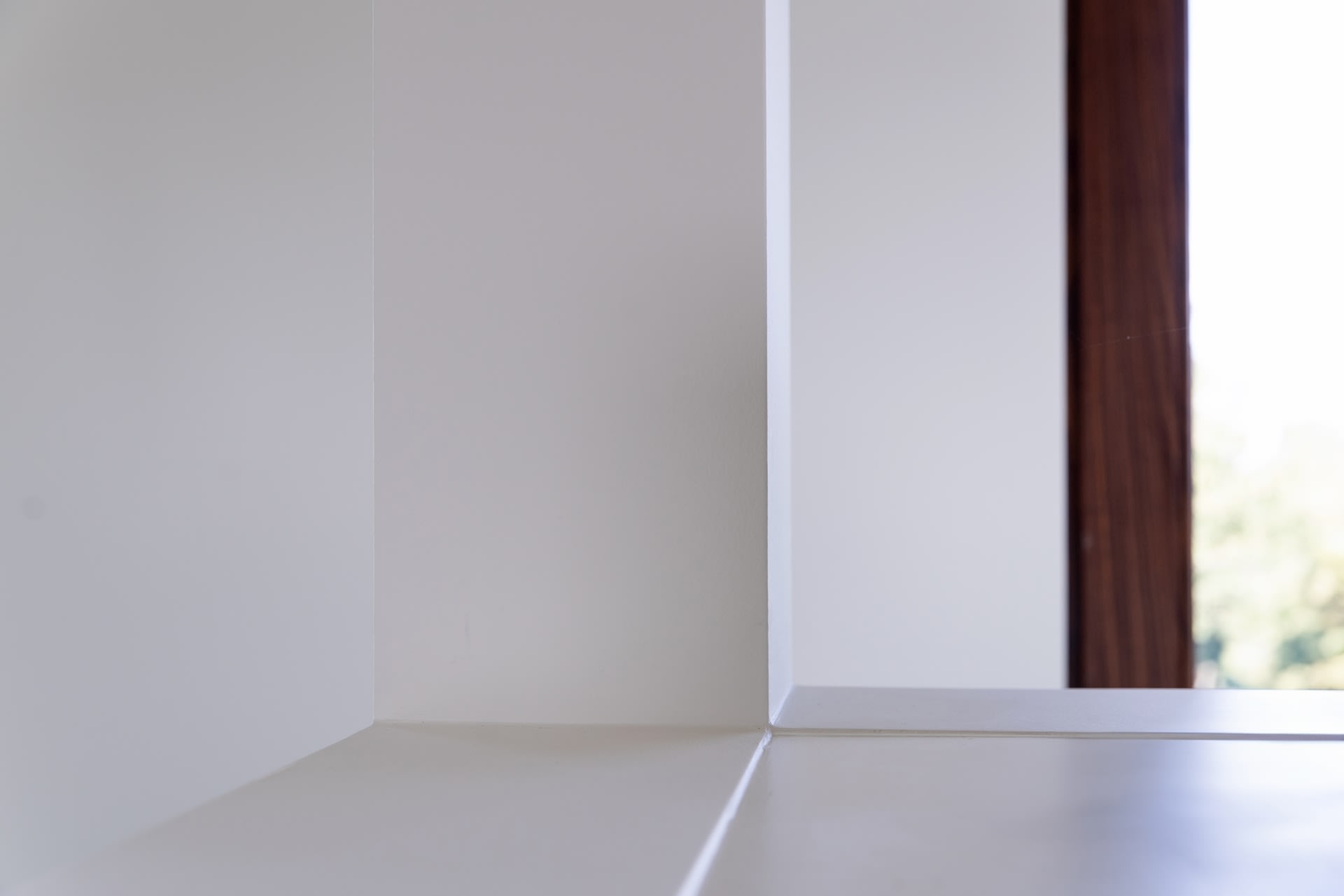 Un coin d'un mur intérieur blanc avec, à droite, une fenêtre dont on aperçoit une partie du cadre en bois et un paysage extérieur flou.