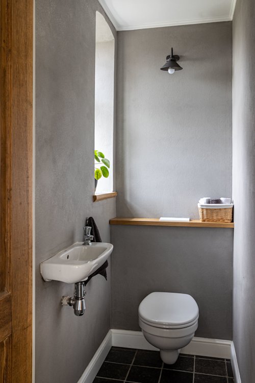 Een kleine badkamer met grijze muren, een toilet aan de muur, een compacte wastafel, een houten plank met een rieten mand en een wandlamp erboven.
