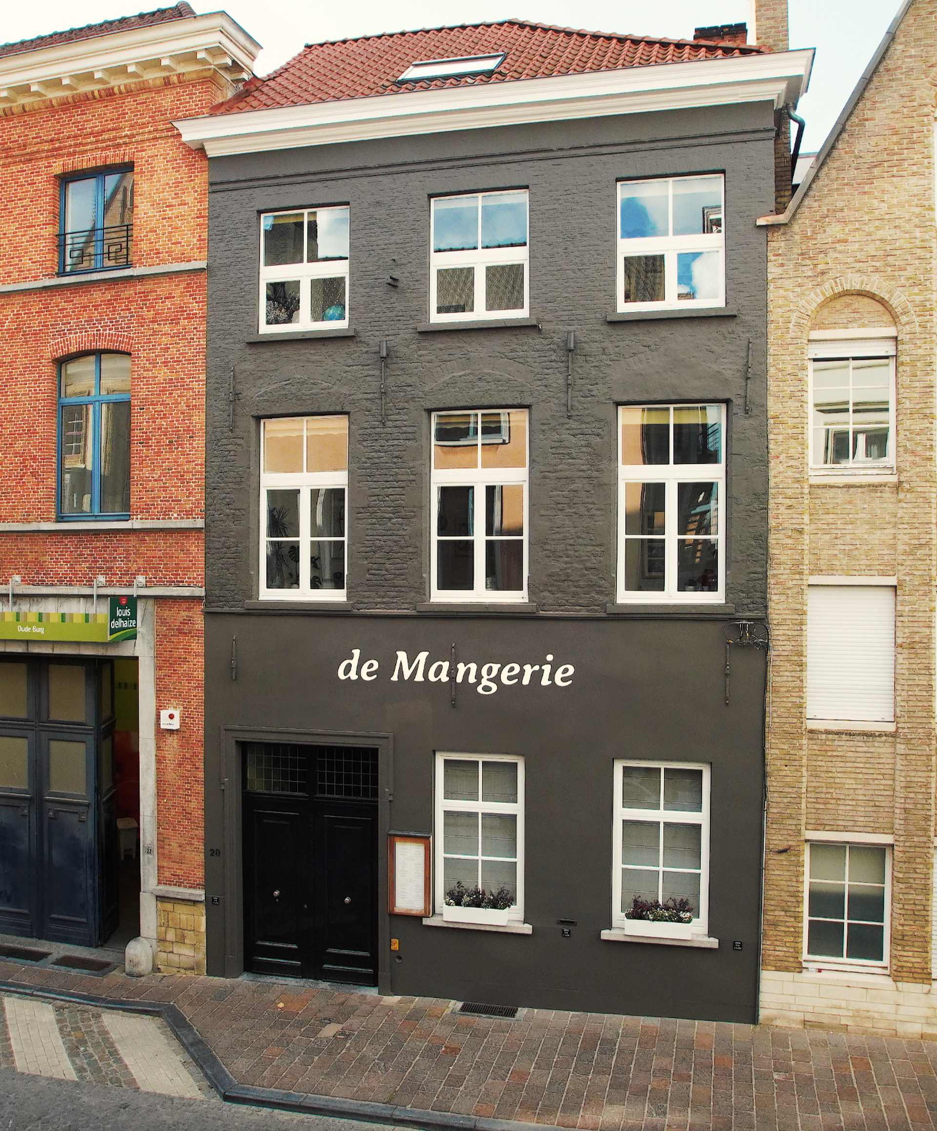 Un bâtiment de trois étages avec une façade grise et les mots « de Mangerie » inscrits sur le devant, situé entre des bâtiments en briques dans une rue pavée.
