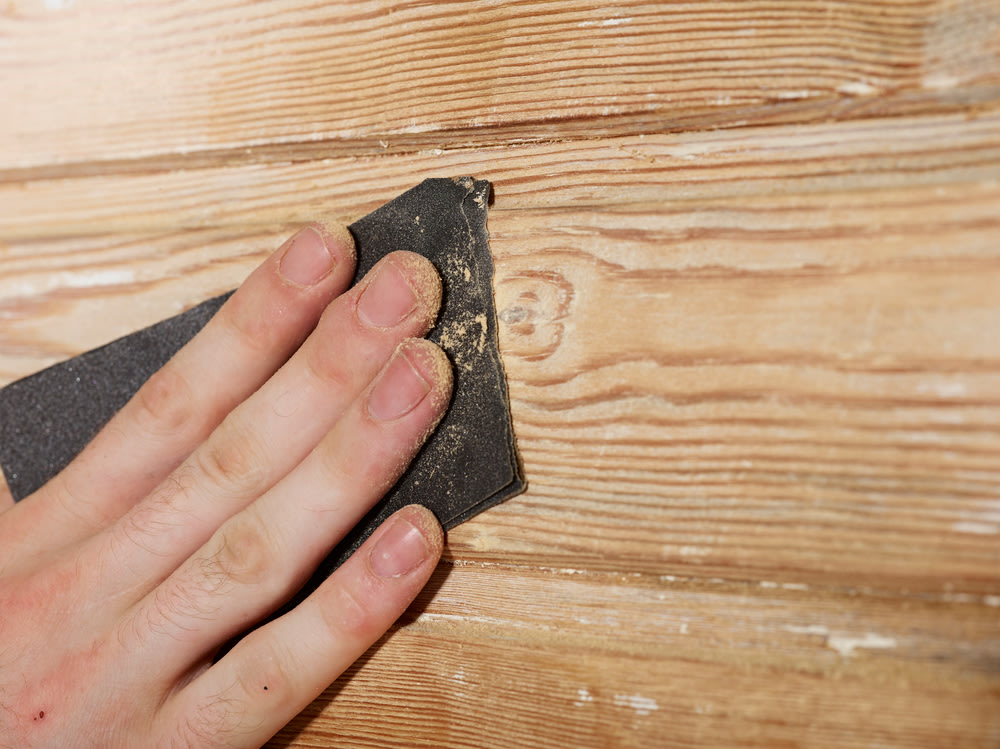 Gros plan sur une main tenant du papier de verre et ponçant une surface en bois, sur laquelle on distingue le grain du bois et un peu de sciure.