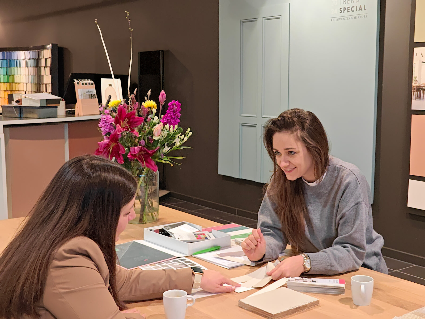 Beeld van kleuradviseur colora en klant aan tafel, ze bespreken de mogelijkheden in je interieur aan de hand van een kleurenkaart, behangstaal, kop koffie tijdens een Interieur Brainstorm Sessie in colora Tielt