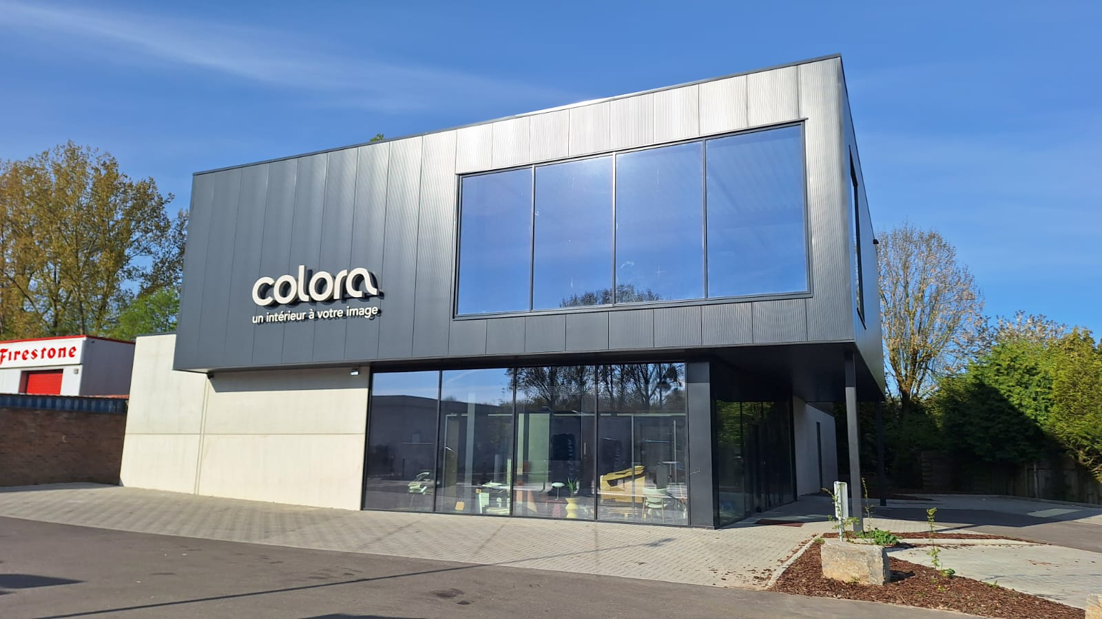 Modern gebouw van twee verdiepingen met grote glazen ramen, grijze buitenpanelen en het "Colora"-logo op de gevel. Het terrein aan de voorkant is bestraat en er staan bomen en groen op de achtergrond.