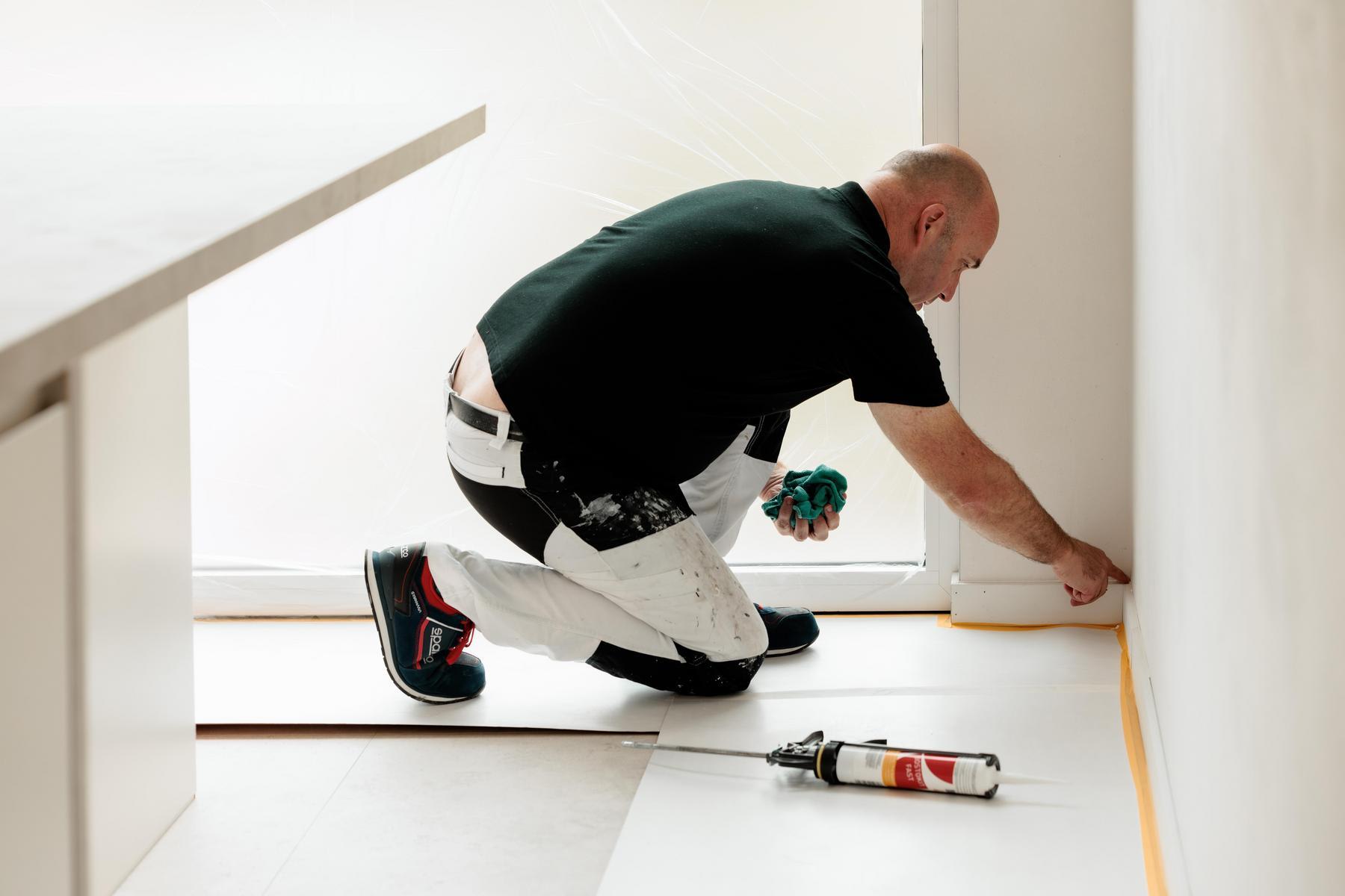 Een man in werkkledij knielt op de grond en wrijft zijn kit aan met de vinger langs de onderkant van een witte muur. Hij werkt voorzichtig in een lichte, moderne kamer.