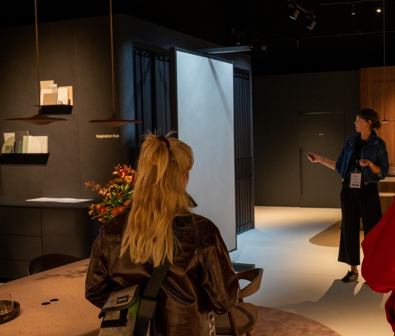 Een persoon met blond haar in een leren jas staat in een showroom terwijl een presentator bij een scherm staat te spreken.