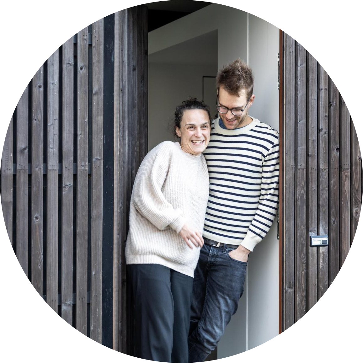 Deux personnes se tiennent ensemble dans l'embrasure d'une porte, l'une vêtue d'un pull couleur crème et d'un pantalon noir, l'autre d'un pull rayé et d'un jean. Toutes deux semblent détendues et souriantes.