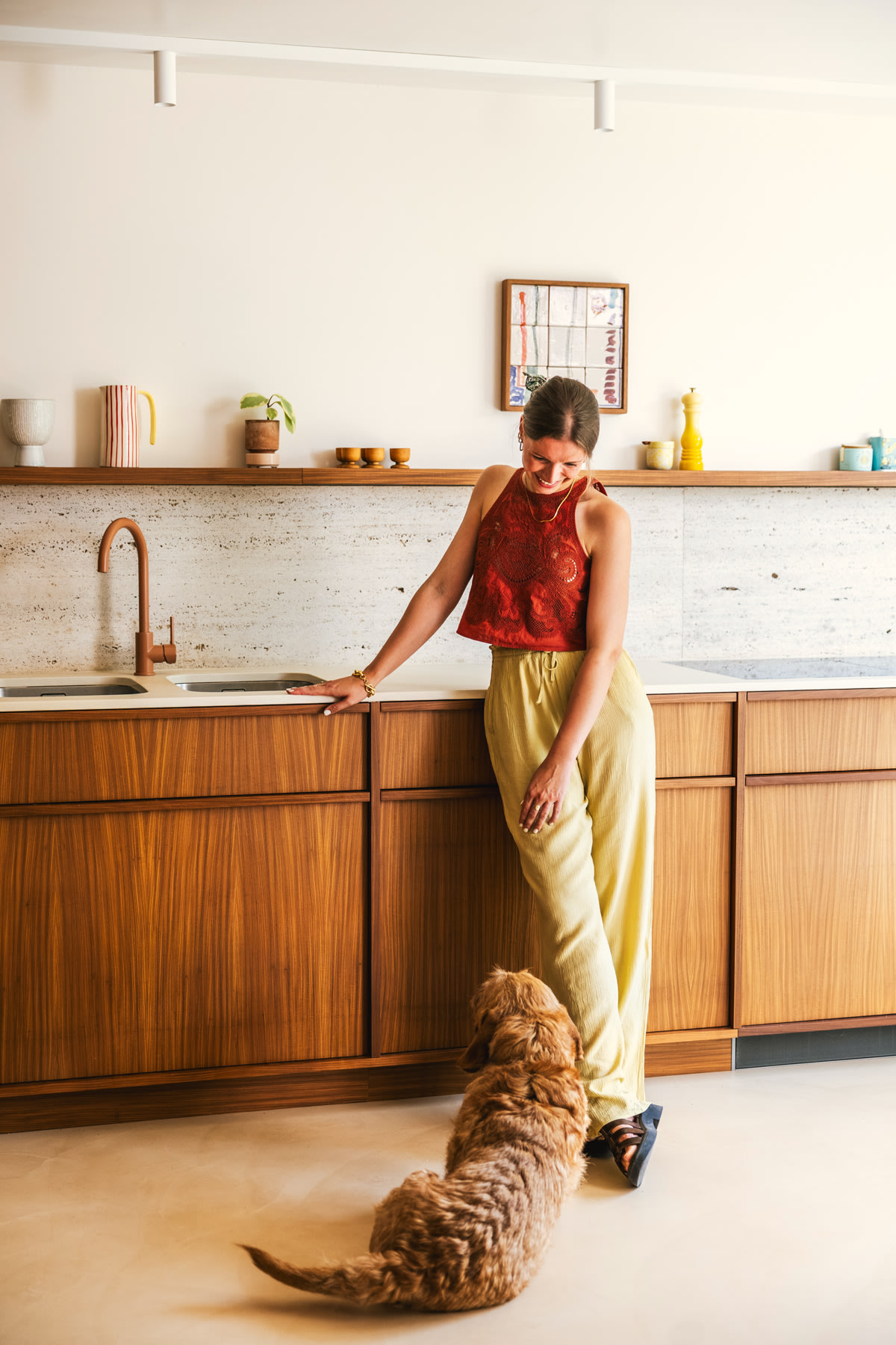 Een vrouw in een rode mouwloze top en gele broek staat in een moderne keuken en glimlacht naar een bruine, pluizige hond die op de grond bij haar voeten ligt. De keuken heeft houten kastjes en een minimale inrichting.