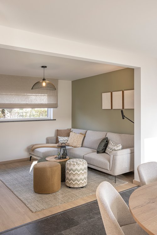 Moderne woonkamer met een beige sofa, ronde poefs, neutrale inrichting, een wandlamp en een raam met een stoffen kap.