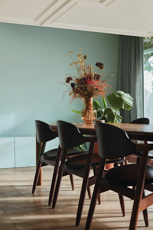 Une salle à manger avec une table en bois, des chaises noires, un vase avec des fleurs séchées et une grande plante verte contre un mur bleu clair baigné de lumière naturelle.