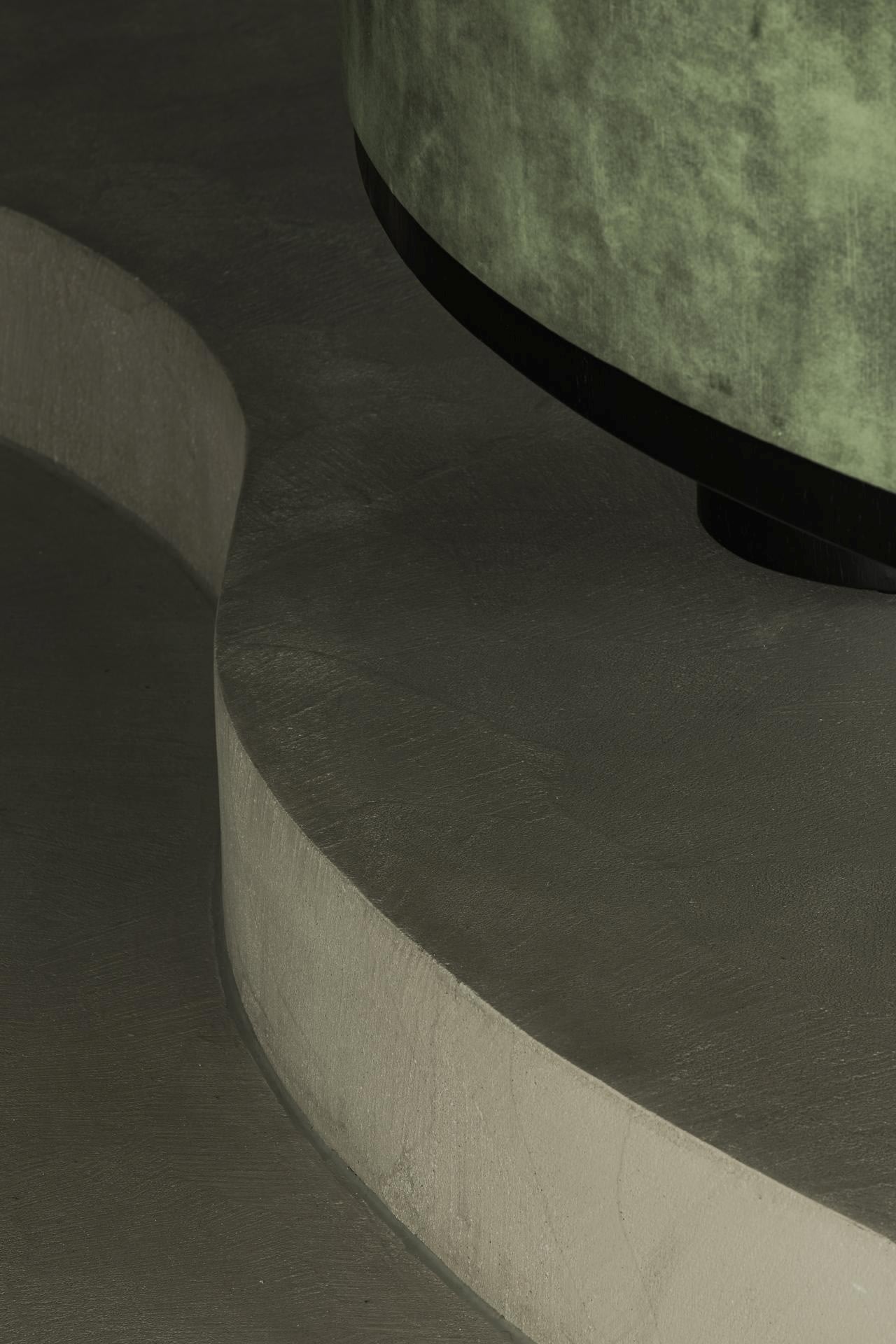 Abstracte weergave van een gebogen betonnen of stenen platform met daarop een gedeeltelijk groen cilindrisch object met textuur, waardoor een minimalistische en moderne compositie ontstaat met zachte verlichting en gedempte kleuren.
