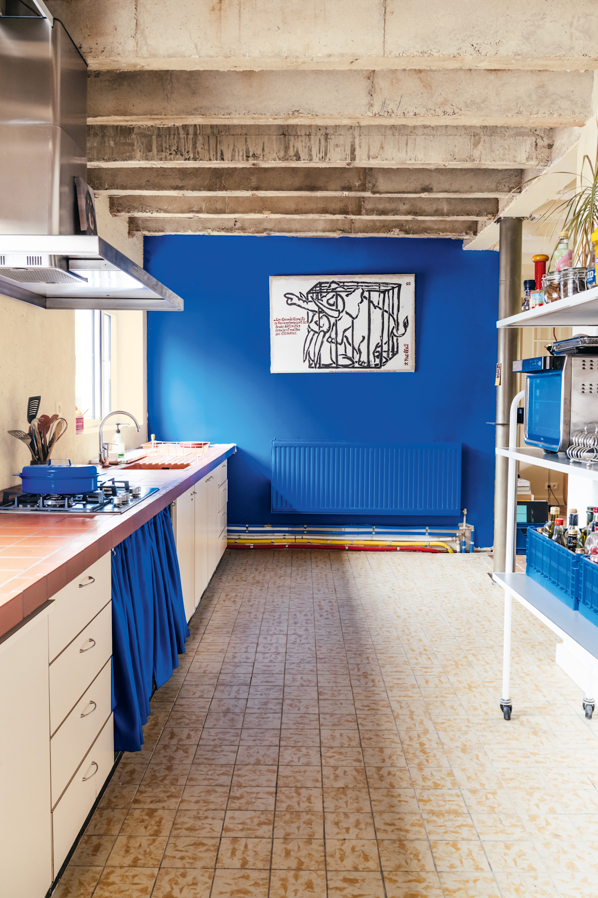 Een keuken met tegelvloer, crèmekleurige kastjes, een blauw gordijn onder het aanrecht, zichtbare plafondbalken en een felblauwe accentmuur met zwart-witte kunstwerken.