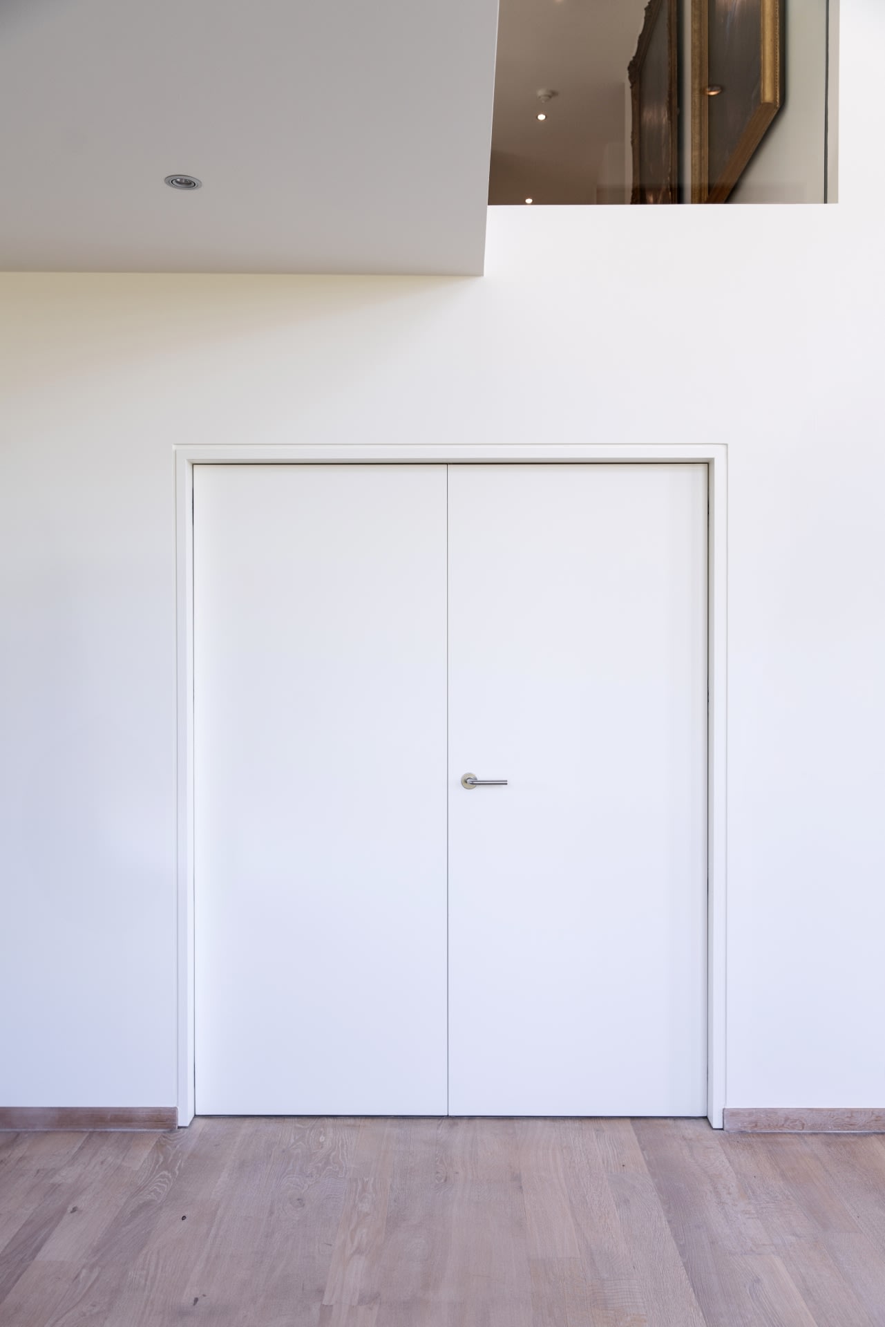 Une double porte blanche fermée avec une poignée argentée est encastrée dans un mur blanc uni, avec un sol en bois clair et une vue partielle sur un étage supérieur.
