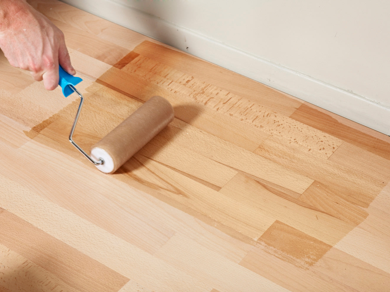 Een hand gebruikt een verfroller om een blanke lak aan te brengen op een houten vloer bij een witte muur.