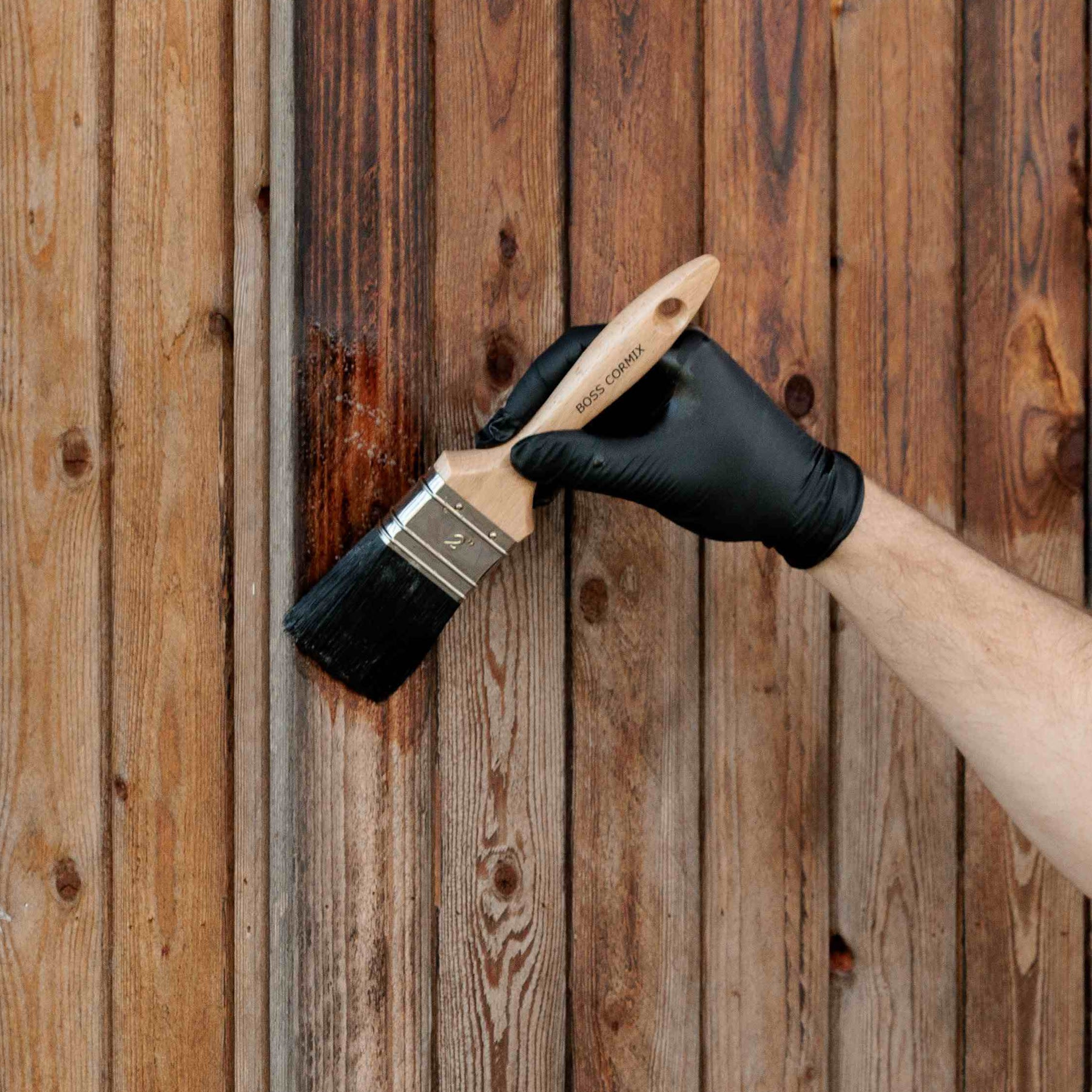 Une personne portant un gant noir applique, à l'aide d'un pinceau, une teinture ou un vernis foncé sur des planches de bois verticales, ce qui met en valeur la couleur et l'aspect du bois.