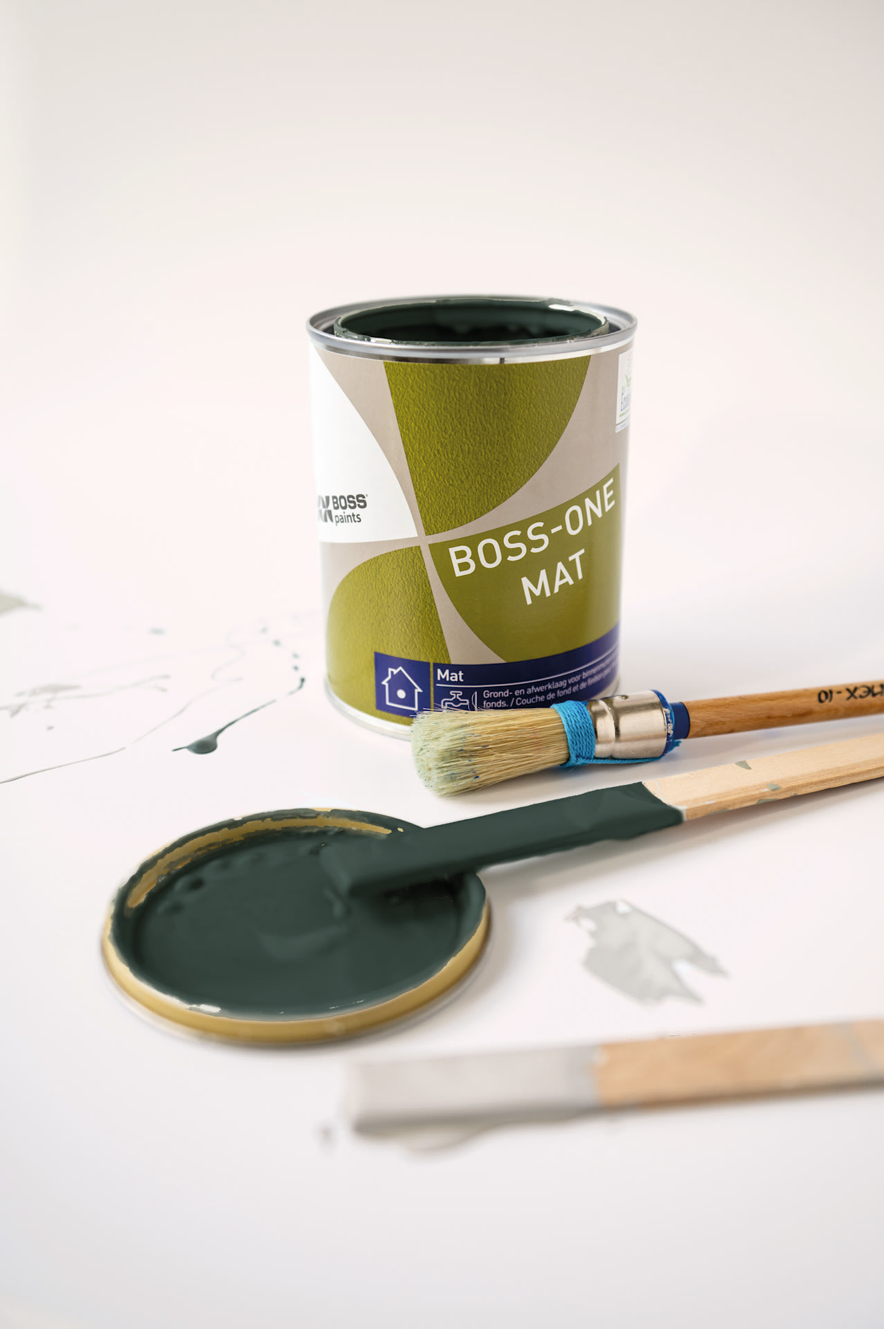 Een blik groene BOSS-ONE MAT-verf staat op een wit oppervlak met het deksel eraf, waardoor de donkergroene verf zichtbaar is. Er liggen twee verfkwasten en er zijn verfspatten in de buurt.