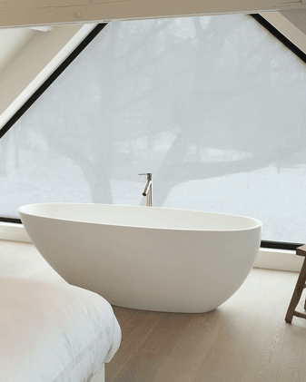 Baignoire blanche îlot devant une grande fenêtre inclinée avec vue sur les arbres, dans une salle de bains lumineuse au sol en bois et aux tons naturels et doux.