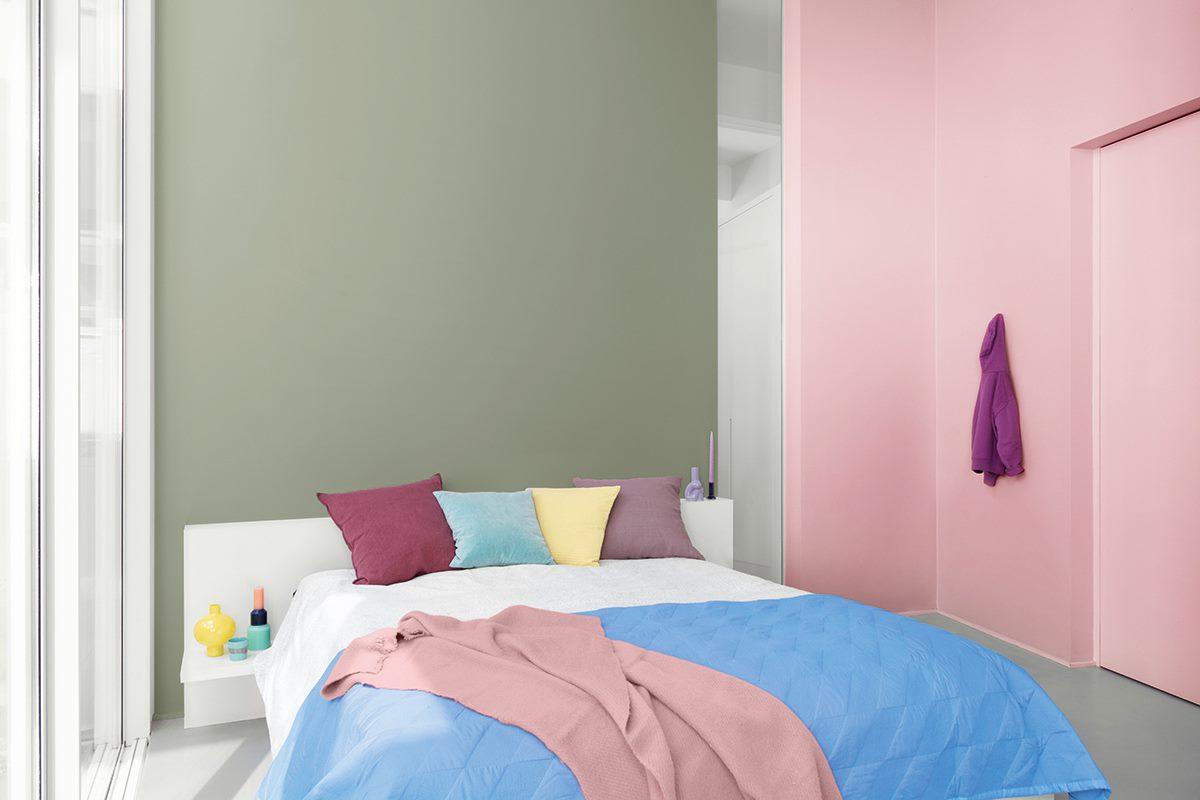 Een moderne slaapkamer met een wit bed, kleurrijke kussens en een blauwe deken. De muren zijn groen en roze geverfd en aan de roze muur hangt een paarse badjas. Licht stroomt door grote ramen.