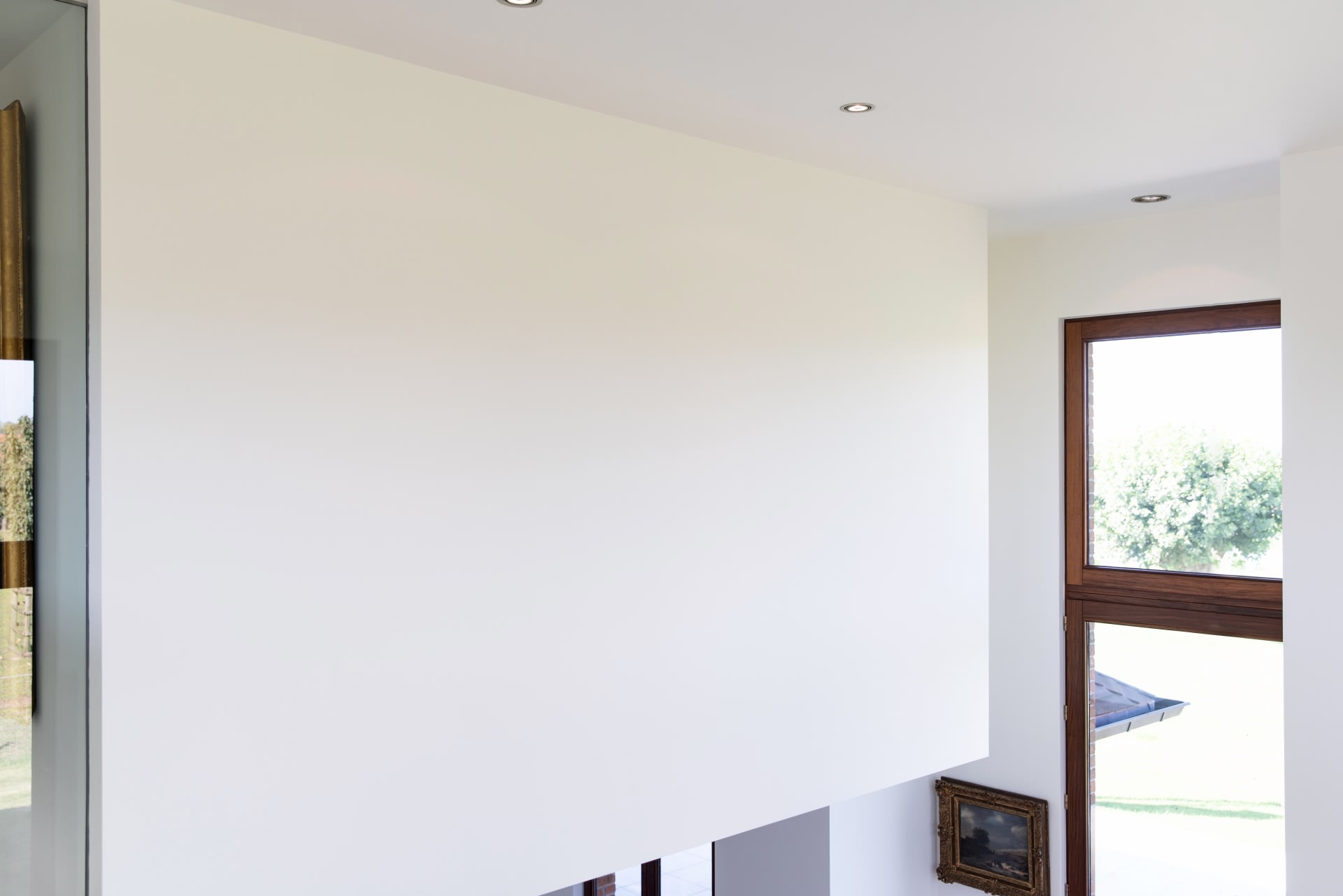Un mur blanc dans un intérieur moderne avec un éclairage encastré au plafond, une fenêtre avec des cadres en bois et des photos encadrées appuyées contre le mur.