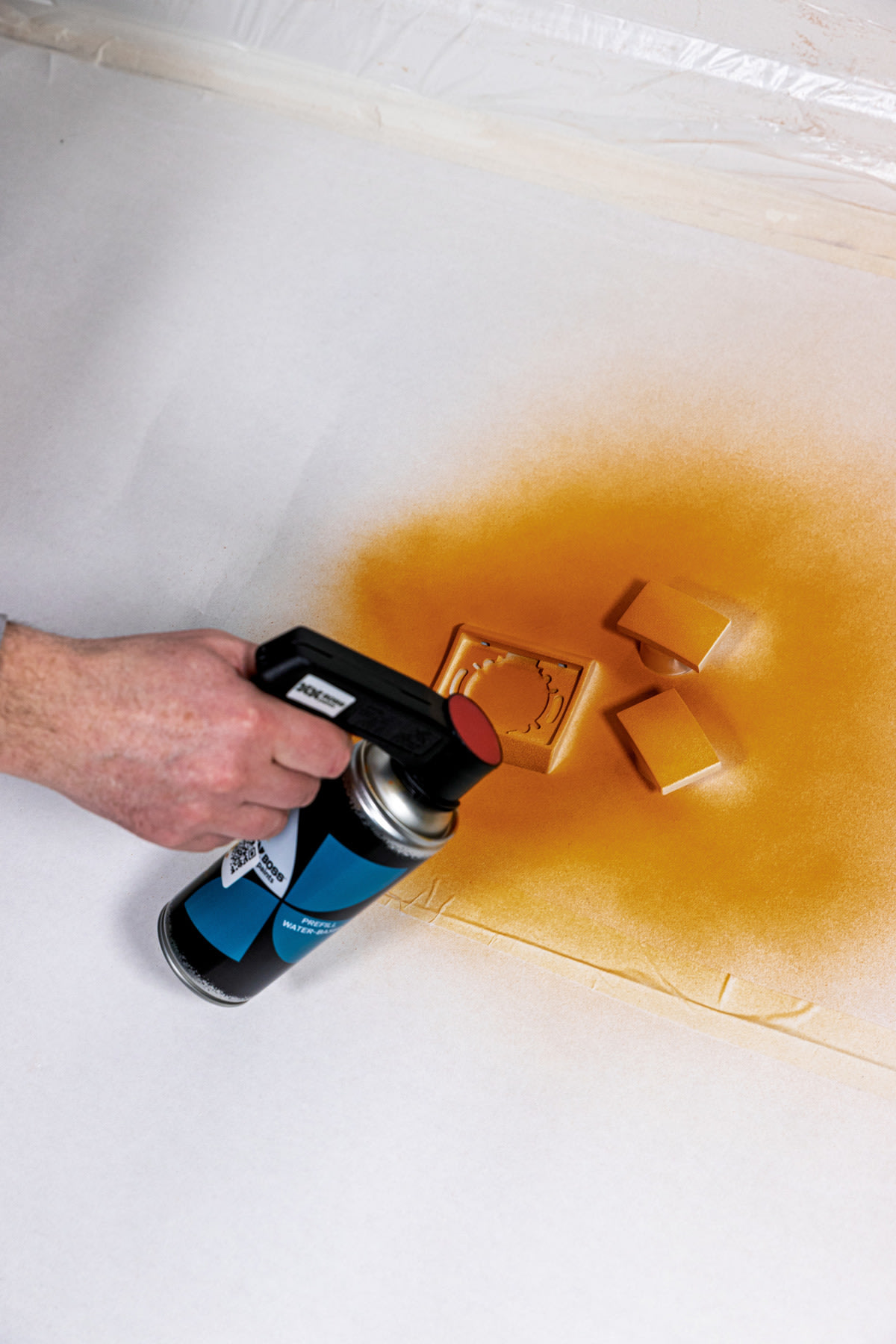 Een hand gebruikt een spuitbus verf om drie kleine rechthoekige voorwerpen en een vierkant voorwerp van oranje verf te voorzien op een wit oppervlak.