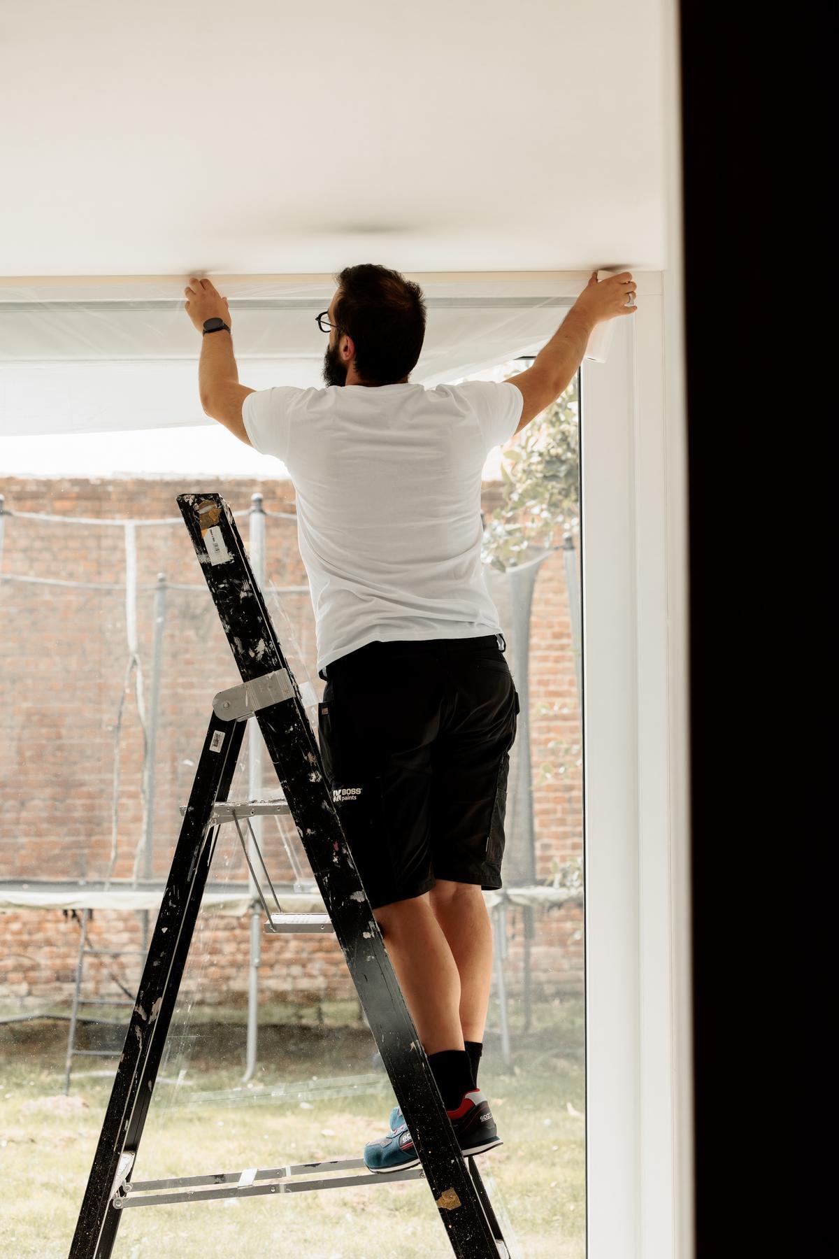 Een man die op een ladder staat, plakt een raam af waarbij door het grote glazen raam de achtertuin en een bakstenen muur te zien zijn.