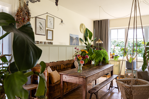 Un coin repas avec une table et des bancs rustiques en bois, décoré de plantes d'intérieur, d'un vase avec des fleurs, d'œuvres d'art encadrées et baigné de lumière naturelle provenant d'une grande fenêtre.