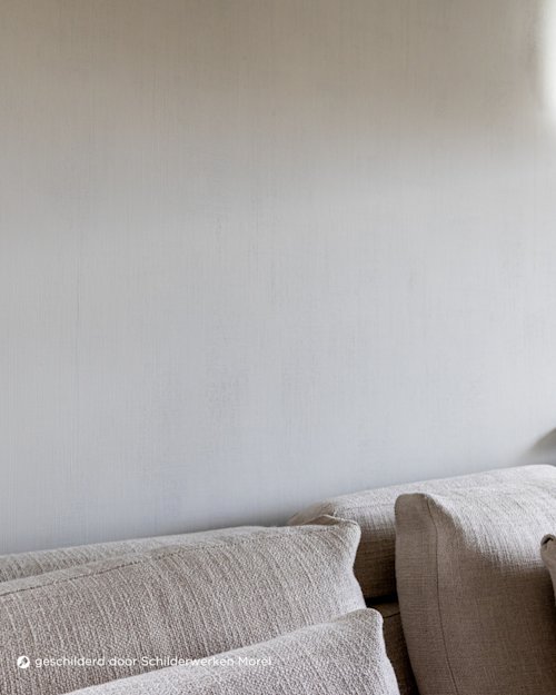 Een close-up van een beige gestructureerde sofa met kussens tegen een effen lichtgrijze muur, gedeeltelijk verlicht door zacht natuurlijk licht van rechtsboven.