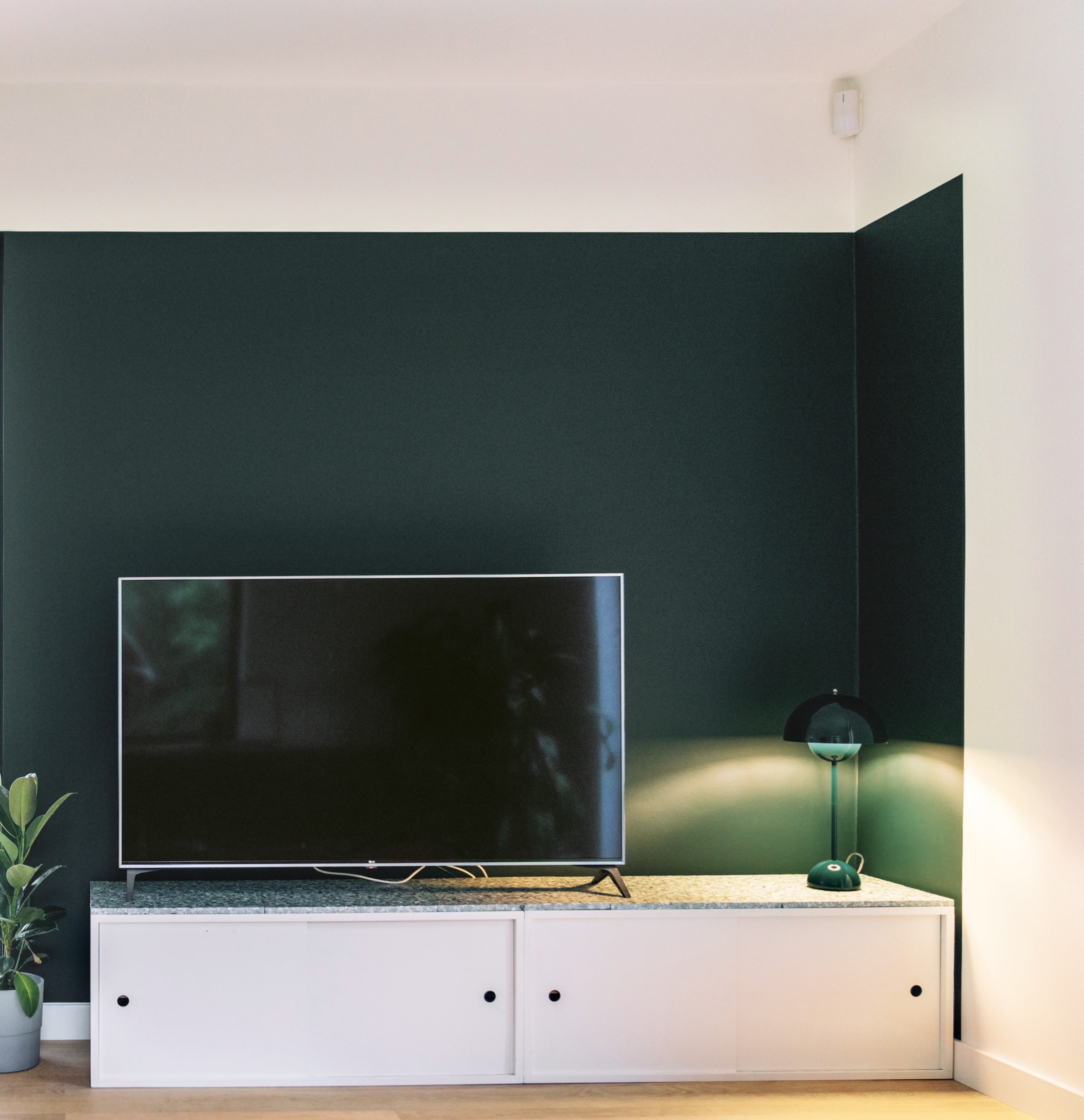 Moderne woonkamer met een tv op een lage witte kast, een groene lamp en een potplant.