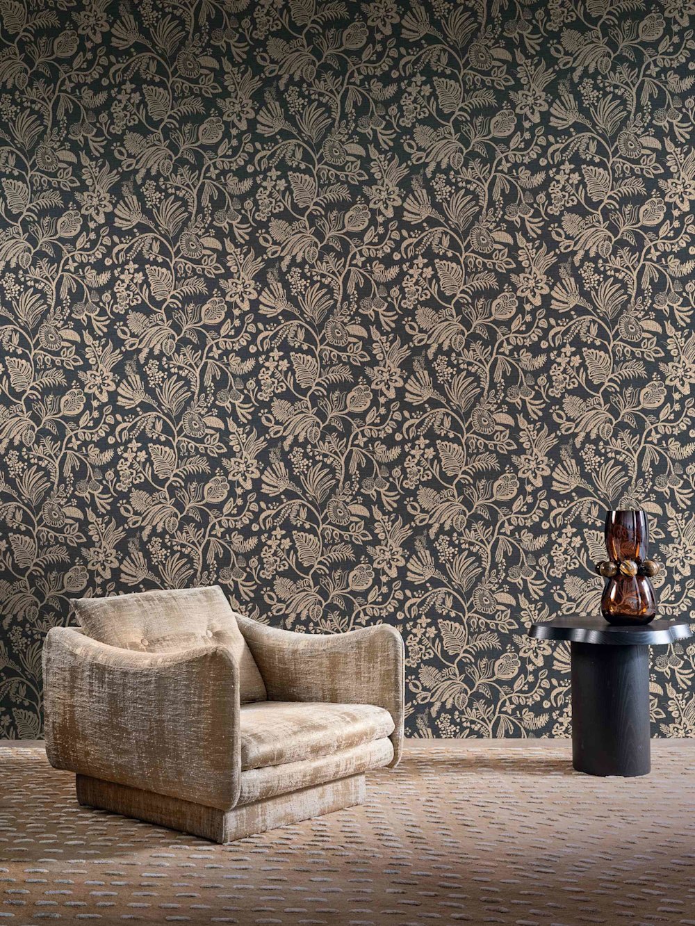 behang - woonkamer - Casamance - Broderies - 76961018  - bruin - zwart- Behang met gouden bloemen op donkere achtergrond, beige fluwelen fauteuil, zwarte bijzettafel, glazen vaas in amberkleur.