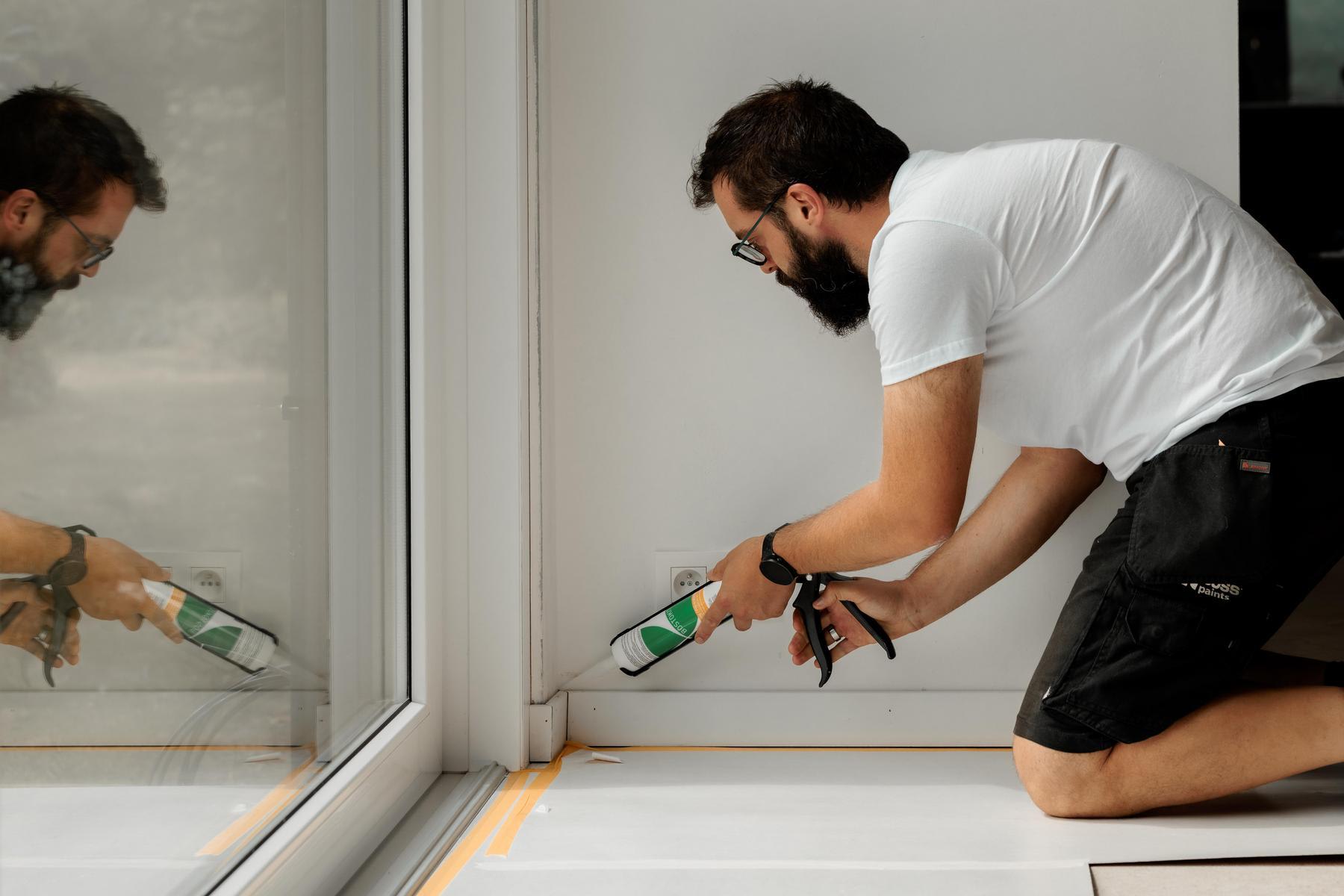 Een man brengt witte kit aan langs de rand van een glazen deurkozijn met een kitpistool.