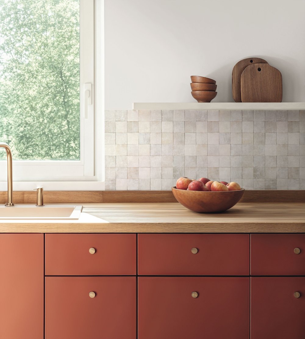 papier peint - cuisine - Caselio - Kitchen box -  KBO106970155 - blanc - Cuisine avec armoires en terre cuite, carrelage mural couleur sable, plan de travail en bois, corbeille à fruits avec des pommes.