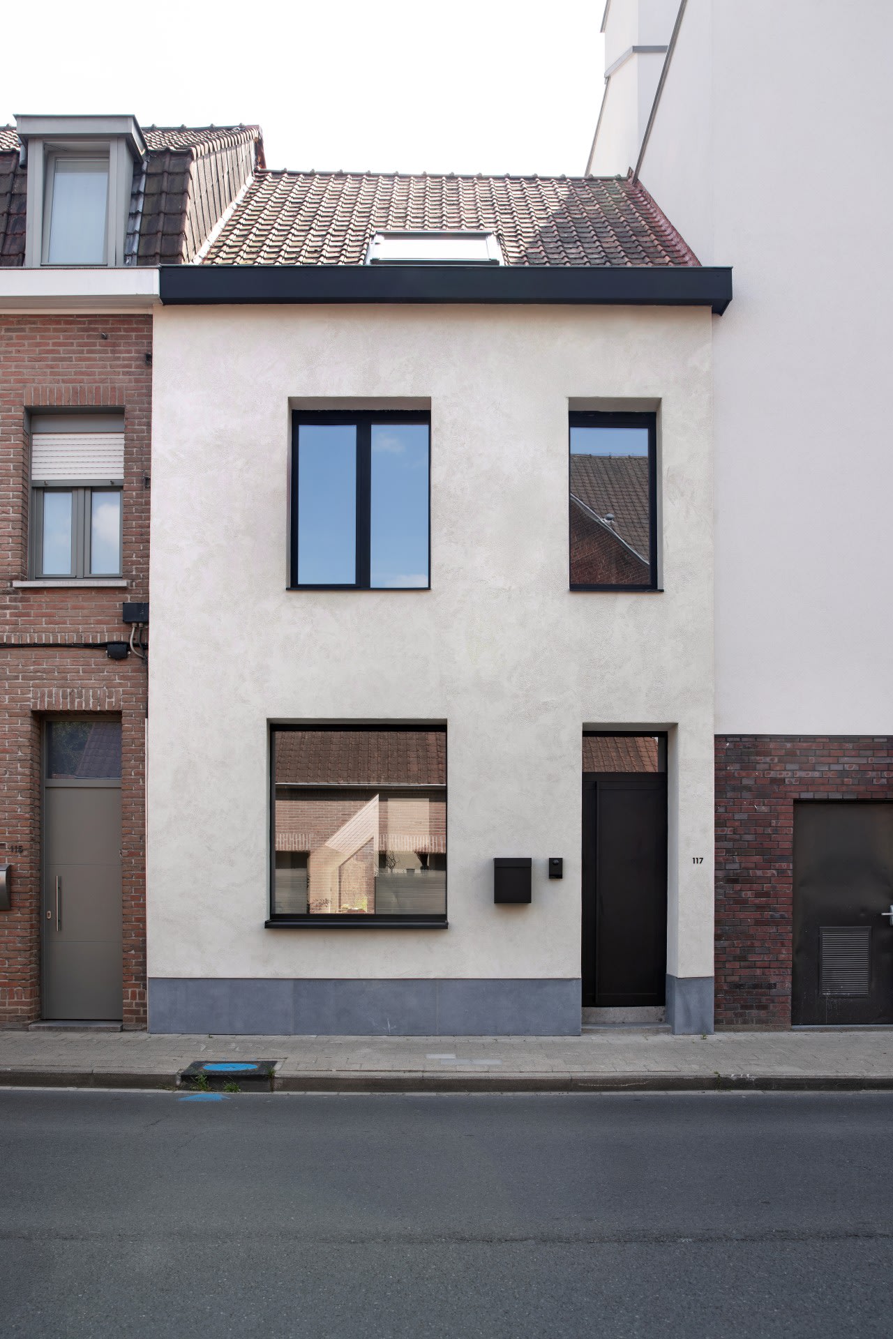 Een huis van twee verdiepingen met een lichtgekleurde stucgevel, ramen met zwarte kozijnen en een donkere toegangsdeur, gelegen tussen twee bakstenen gebouwen in een stadsstraat.