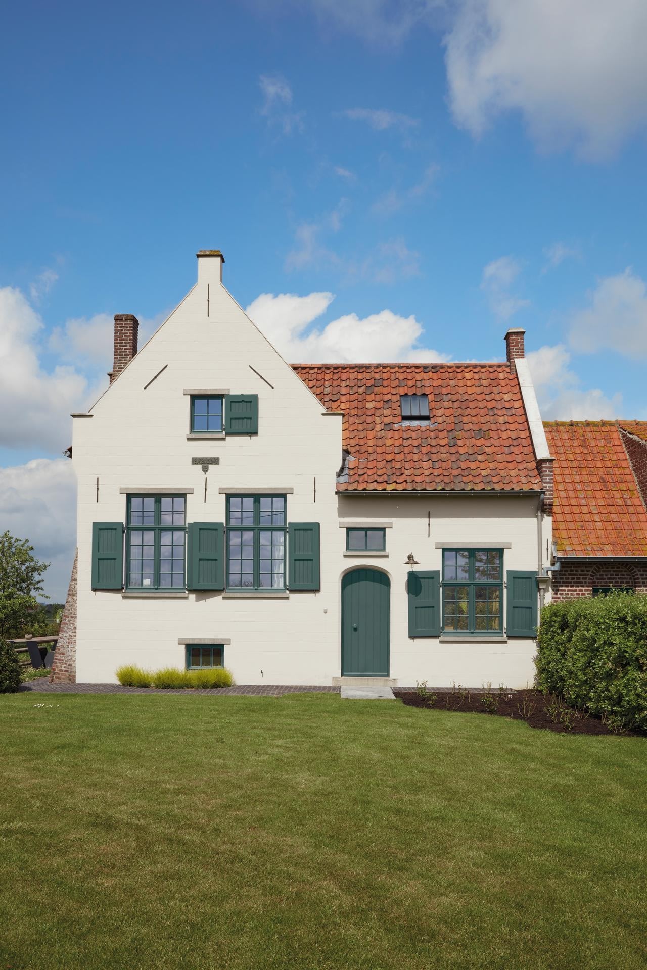 Een wit huis met groene luiken en een rood pannendak staat op een netjes getrimd gazon onder een blauwe lucht met verspreide wolken. Struiken en heesters omzomen de tuin.