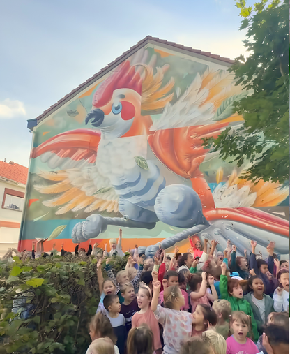 Een grote, kleurrijke muurschildering van een vogel siert de muur van een gebouw. Daarvoor staat een groep opgewonden kinderen buiten; sommigen hebben hun handen omhoog, glimlachen of juichen onder een gedeeltelijk zonnige hemel.