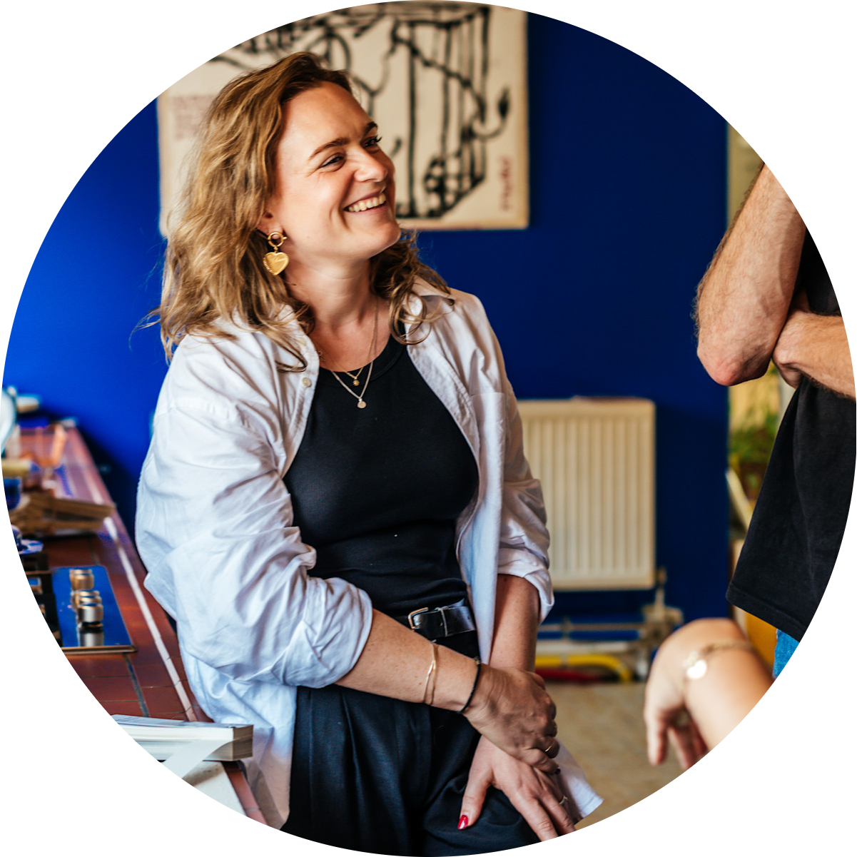 Een vrouw in een wit shirt en een zwarte broek glimlacht terwijl ze tegen een toonbank leunt, in gesprek met twee mensen in een helder verlichte kamer met blauwe muren.