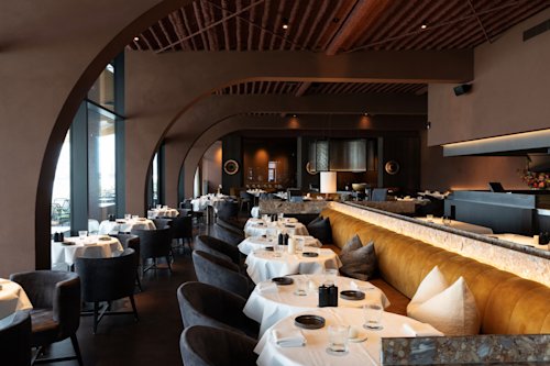 Modern restaurantinterieur met gewelfde muren, donkere stoelen, witte tafelkleden en een lange gestoffeerde bank onder zachte sfeerverlichting.