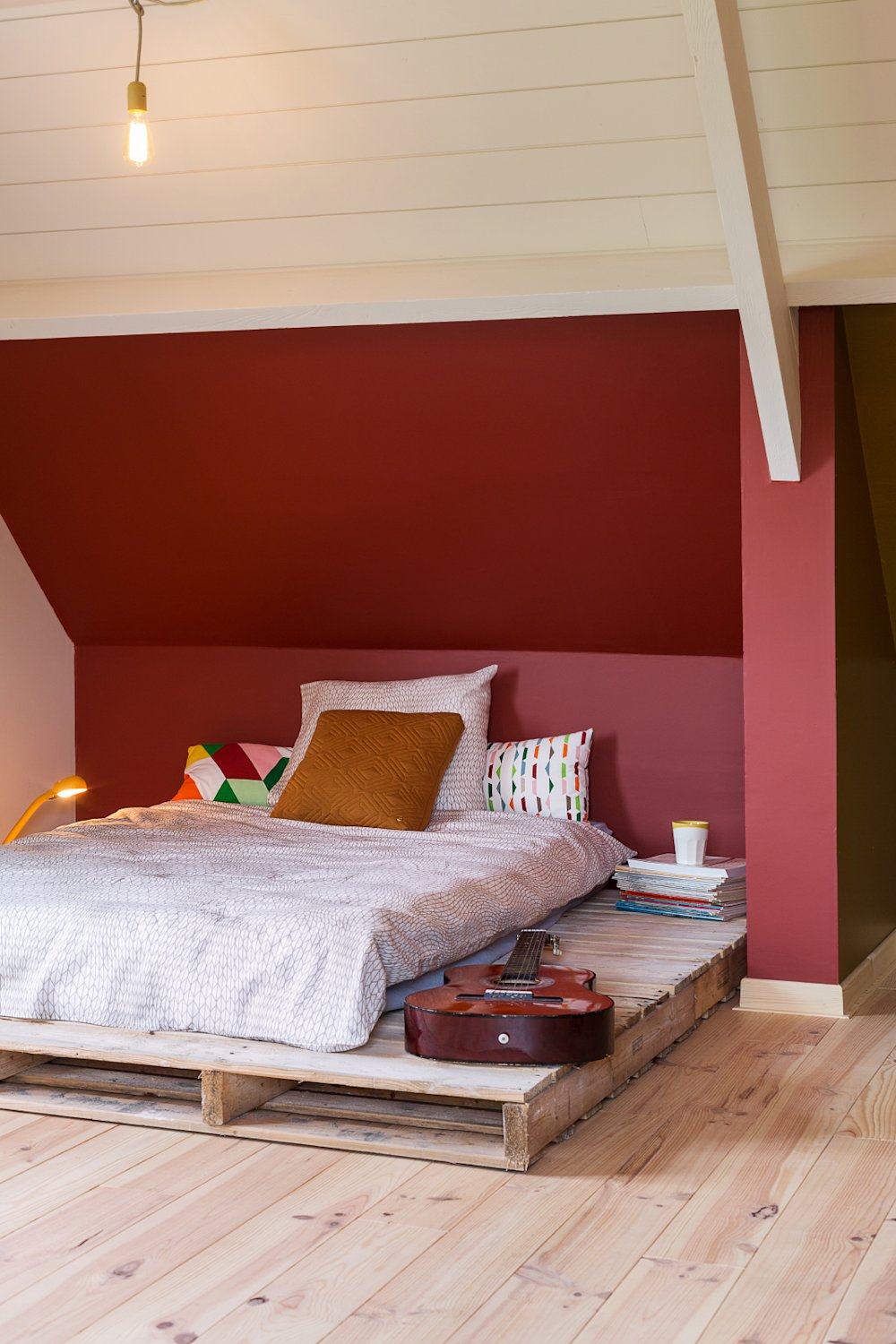 Colora-slaapkamer met warm rode accentmuur achter het bed, witte houten plafondlatten en houten vloer