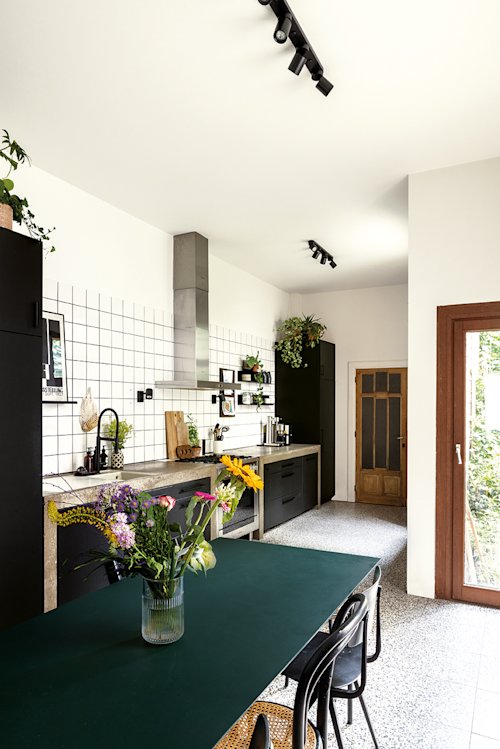 keuken met witte muren en witte tegels, donkergroene eettafel met bloemen, zwarte keukenkasten en houten ramen