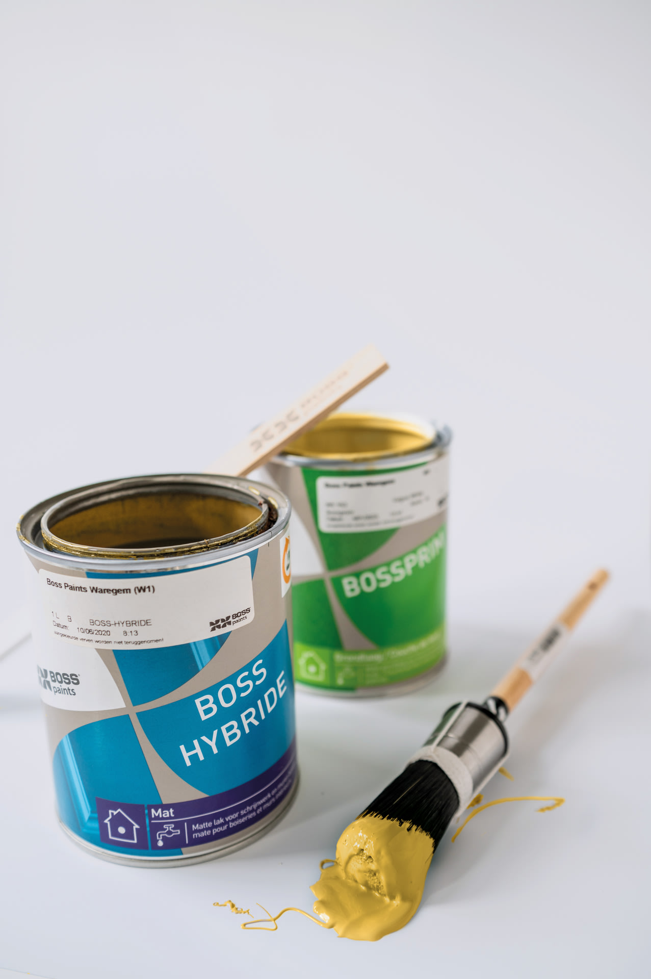 Deux pots de peinture ouverts sont posés sur une surface blanche. Devant les pots se trouve un pinceau recouvert de peinture jaune. Sur l'un des pots repose un petit bâtonnet en bois.