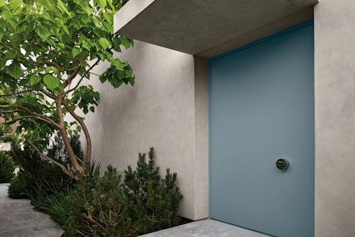 Een moderne blauwe voordeur met een ronde handgreep op een lichtgekleurd huis, omringd door groen en betonnen oppervlakken.