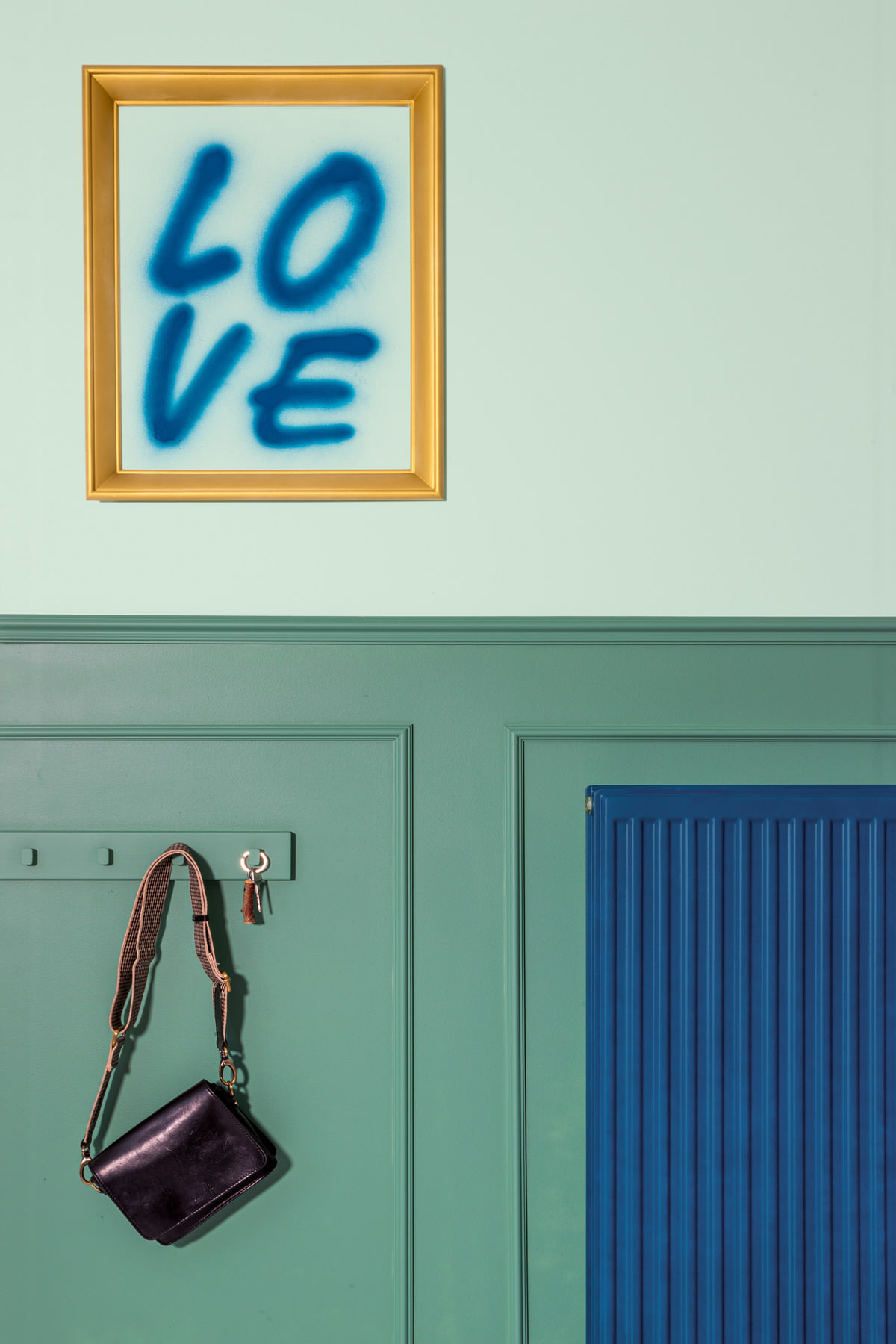 Een blauw gespoten "LOVE" in lijst hangt boven een groene muur met een zwarte tas en sleutels aan een haak naast een blauw radiatorpaneel.