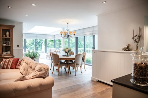 Open woon- en eetruimte met een beige bank, ronde eettafel, kroonluchter, grote ramen en uitzicht op een tuin buiten.