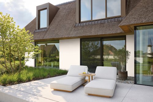 Twee moderne loungestoelen en een tafeltje met drankjes op een patio buiten een modern huis met grote ramen en een rieten dak.