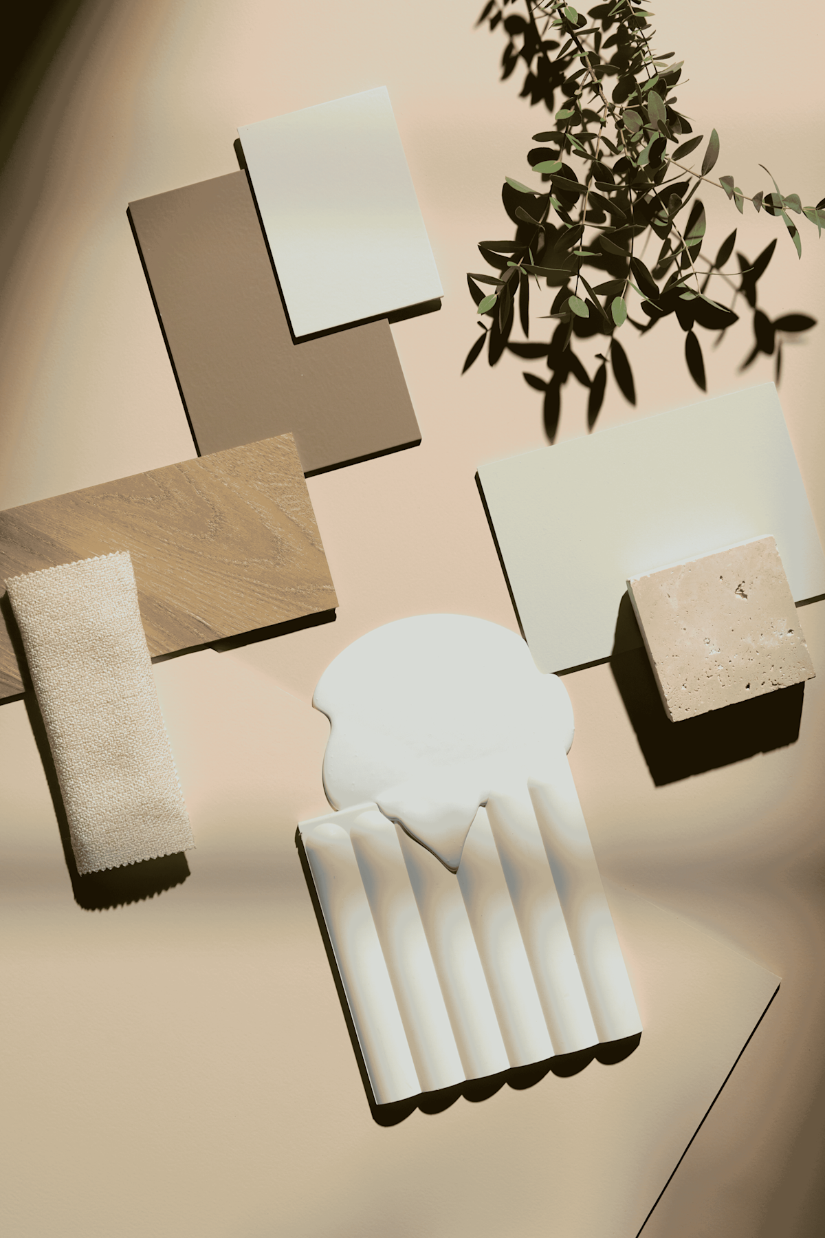 Een ‘flat lay’ van tegels in neutrale tinten, een stofstaal, steen met textuur, een keramisch voorwerp met witte glazuurlaag en een tak die een schaduw werpt, allemaal gerangschikt op een beige ondergrond.
