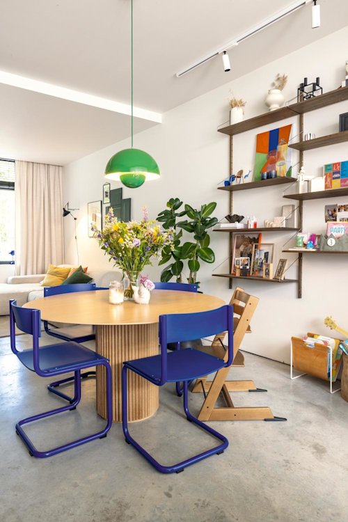 Een moderne eethoek met een ronde houten tafel, vier blauwe stoelen, een groene hanglamp, verse bloemen en wandplanken met boeken en decor.