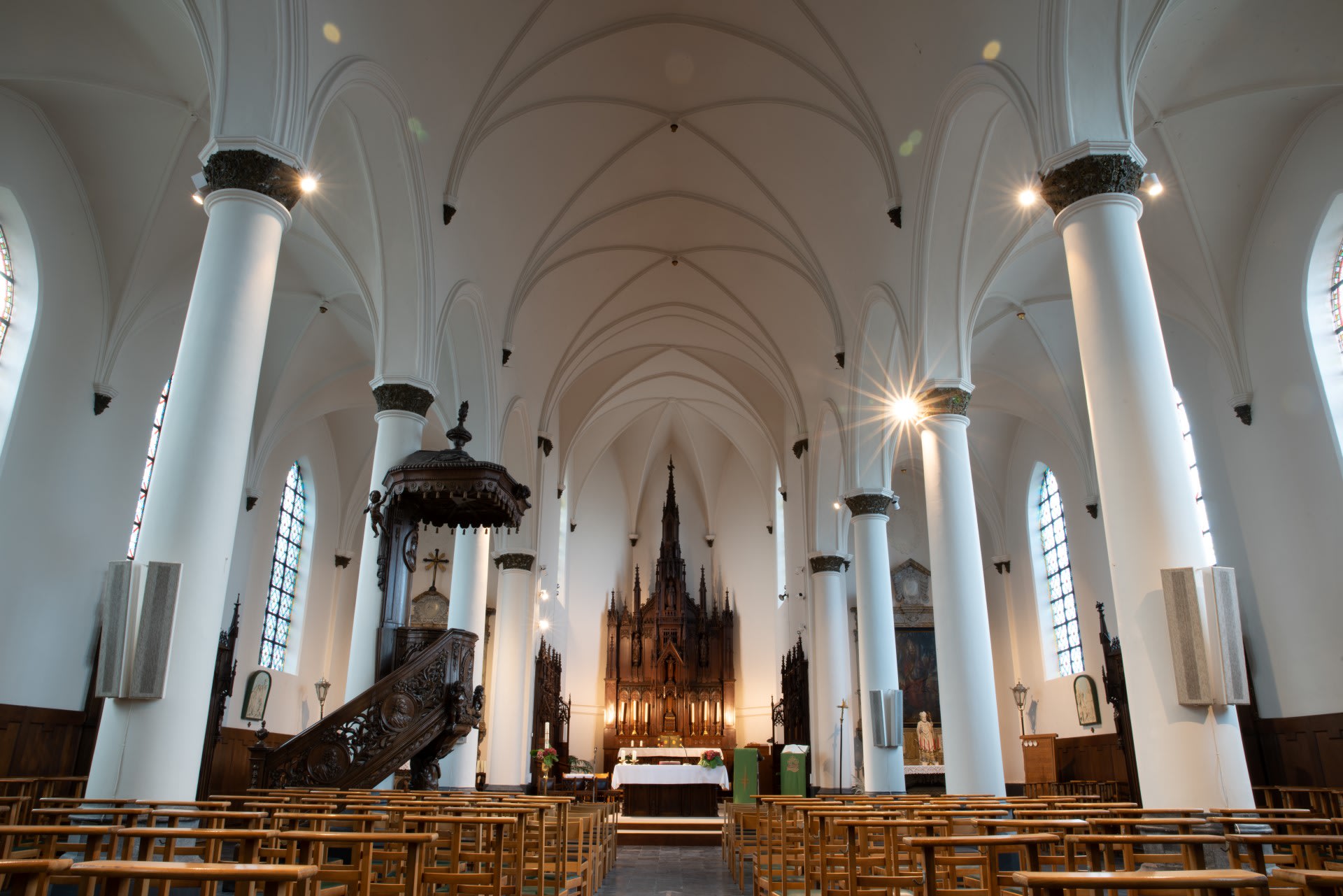 Interieur van een grote kerk met witte gewelfde plafonds, hoge zuilen, houten kerkbanken, glas-in-loodramen en een sierlijk altaar aan de voorkant.