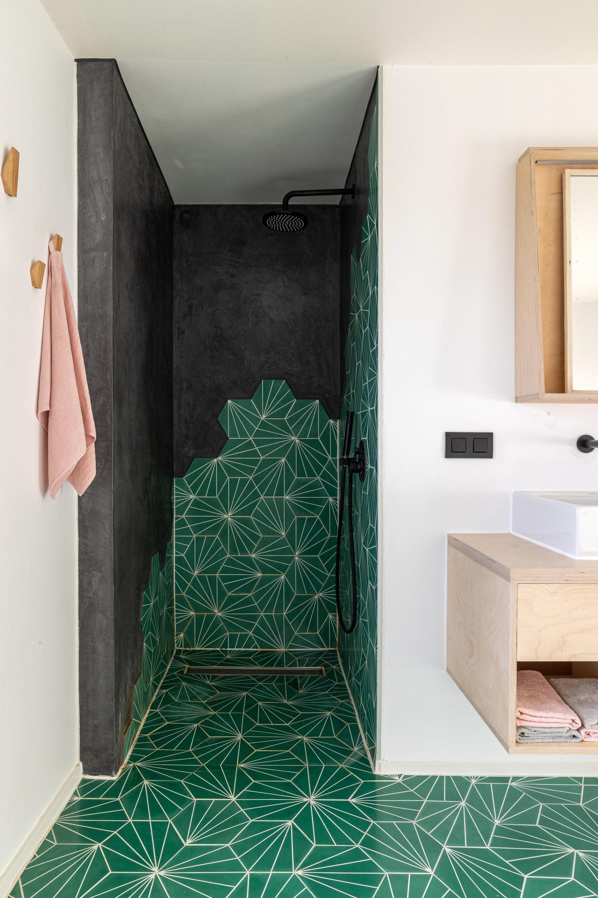 Salle de bain moderne avec douche à l'italienne, murs noirs et motif géométrique vert sur le sol et les murs, meuble de salle de bain en bois et serviette rose accrochée au mur.