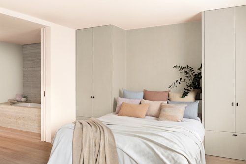 Een moderne slaapkamer met een opgemaakt bed, kussens in neutrale tinten, ingebouwde kasten, een plant op een zijplank en een gedeeltelijk uitzicht op een badkamer met een bad.